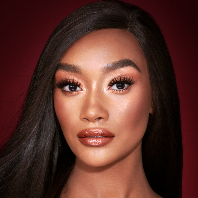 A deep-tone model with brown eyes wearing shimmery copper and gold eye makeup with black eyeliner, glowy bronzed cheeks, and orange-red lipstick with gloss on top. 