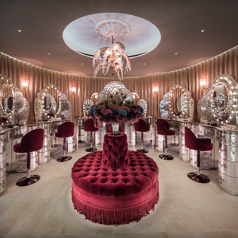 A red and golden-themed make-up store with revolving chairs and mirrored-dressing tables with LED lights. 