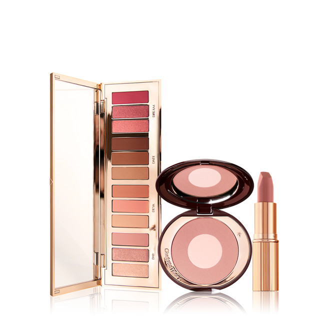 An open, mirrored-lid eyeshadow palette with matte and shimmery nude shades, a two-tone powder blush in nude pink, and a nude pink lipstick in a golden-coloured tube. 