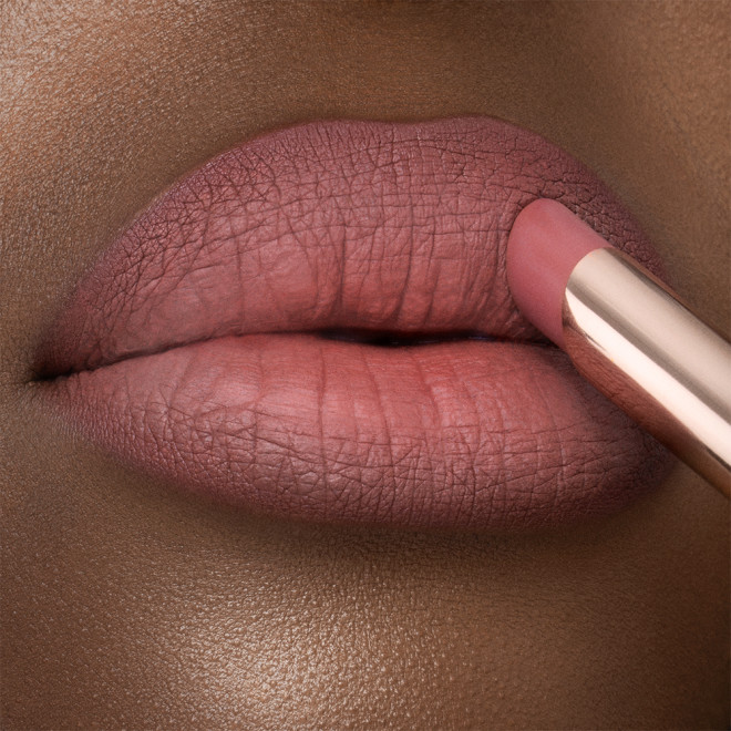 Lips close-up of a deep-tone model applying a lipstick in a neutral rosy-pink colour with a matte finish.