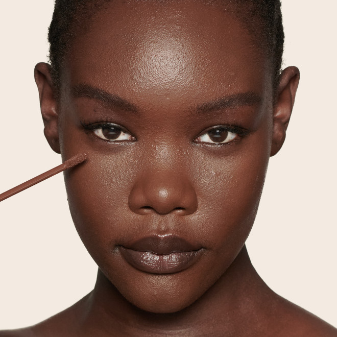 Deep-tone model with brown eyes wearing a radiant, concealer that brightens, covers blemishes, and makes her skin look fresh along with nude lip gloss and subtle eye makeup.