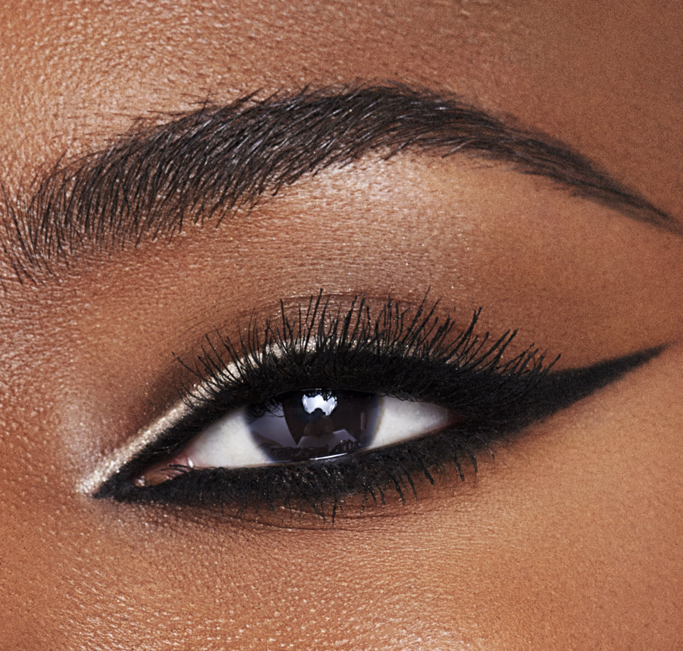 Single eye close-up of a deep-tone model with brown eyes wearing a duo eyeliner in black and champagne-beige on her upper lid and lower waterline. 