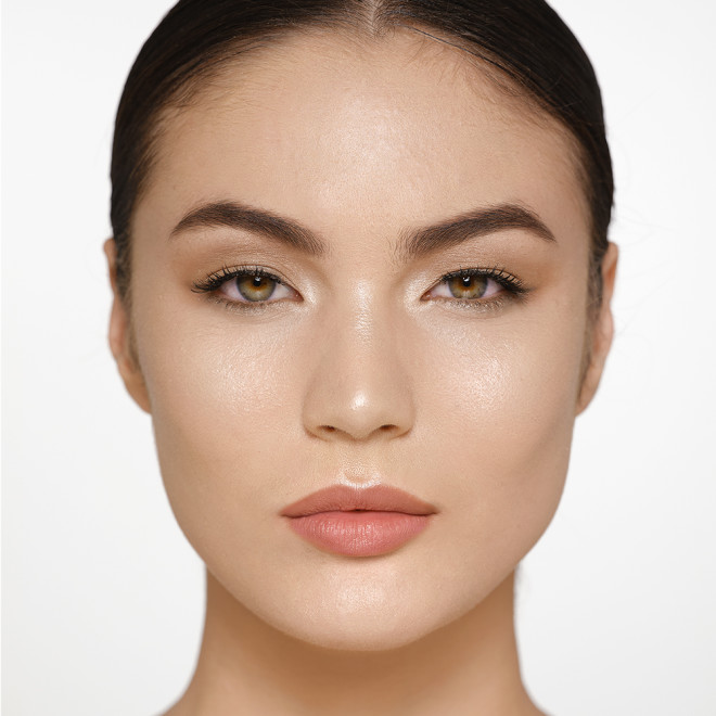 Fair-tone brunette model wearing glowy highlighter in a silvery-gold shade with nude pink lip lipstick and soft eye makeup.