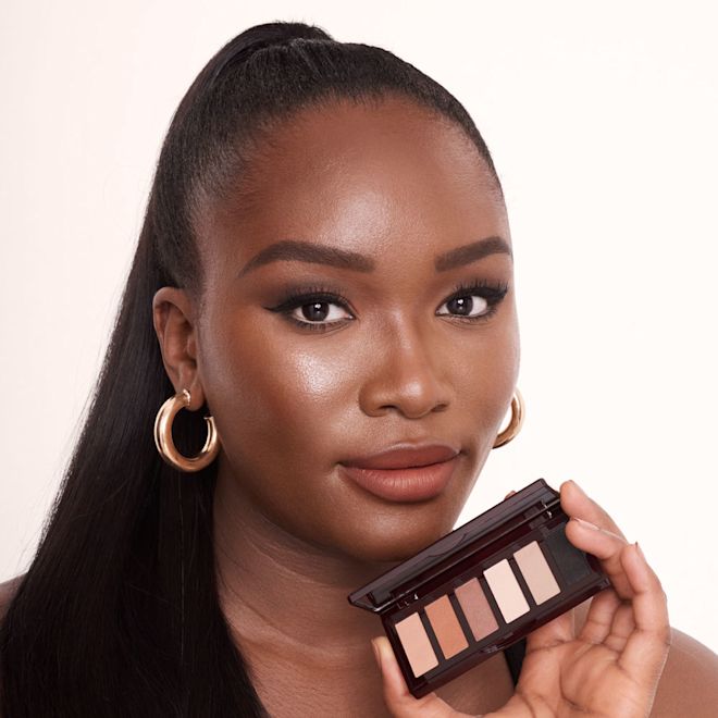Super Nudes Easy Eye Palette on deep-tone model A deep-tone model with brown eyes holding an open, mirrored-lid six-pan eyeshadow palette with matte eyeshadows in brown, peach, and beige shades while wearing a smokey brown eye look created using that palette with nude peach lipstick.