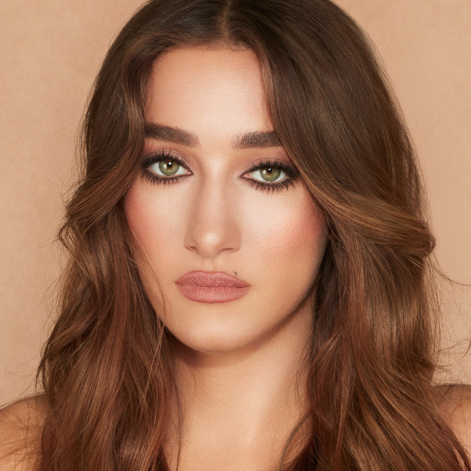 Medium-tone model with green eyes wearing soft beige and champagne eyeshadow and black kohl liner with a fresh, neutral nude peach lipstick with a matte finish.