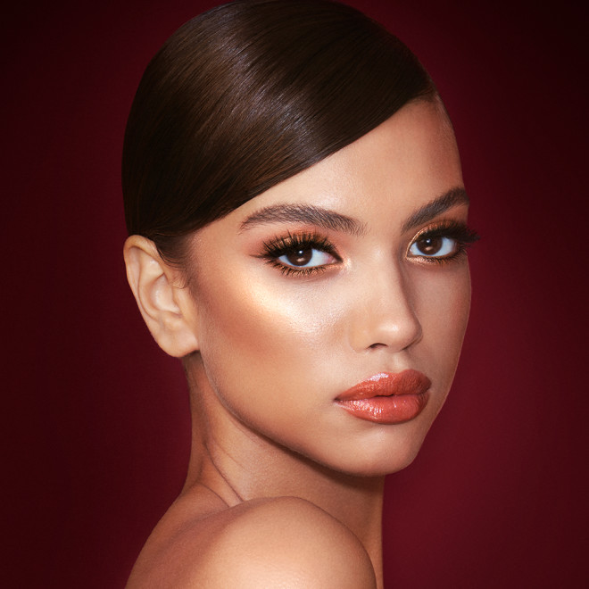 A medium-tone model with brown eyes wearing shimmery copper and gold eye makeup with black eyeliner, glowy bronzed cheeks, and orange-red lipstick with gloss on top. 