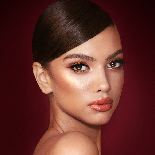The Queen of Glow look on medium-tone model A medium-tone model with brown eyes wearing shimmery copper and gold eye makeup with black eyeliner, glowy bronzed cheeks, and orange-red lipstick with gloss on top.