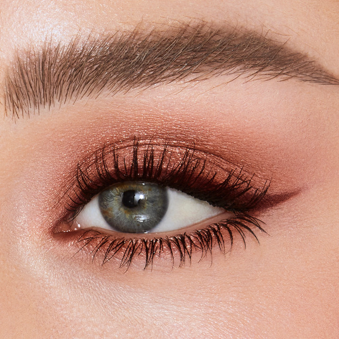 Single-eye close-up of a fair-tone model with blue eyes wearing a shimmery russet rose cream eyeshadow with a golden sparkle and red-brown eyeliner.