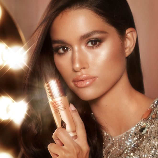 Hollywood Flawless Filter held by medium-tone model Medium-tone model with glowy, flawless skin holding a luminous primer in a medium shade in a glass bottle with a gold-coloured lid.