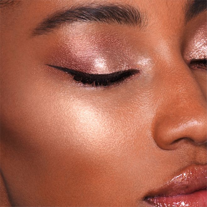 Hollywood Superstar Glow Highlighter on deep-tone model close-up Close-up of a deep-tone model wearing shimmery rose-gold eyeshadow and black eyeliner with a glowy yet subtle, pearlescent highlighter in a rose-gold shade.