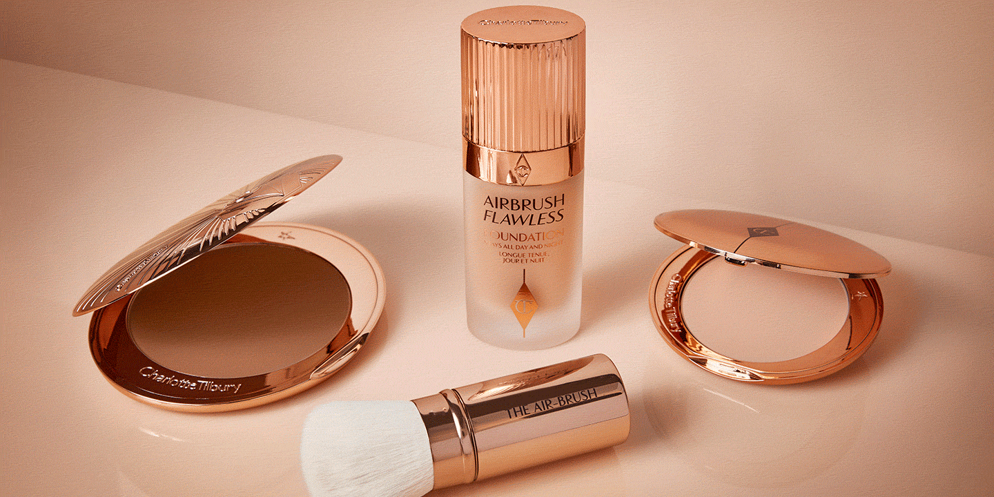 An open pressed powder compact in a light beige shade with a foundation in a frosted glass bottle with a gold-coloured lid, a kabuki brush with a gold-coloured lid and white bristles, and a bronzer compact in gold-coloured packaging.