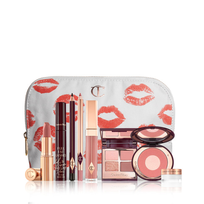 A mascara with an open, mirrored-lid quad eyeshadow palette in shimmery neutral shades, a shimmery nude pink lipstick, a nude pink lip gloss and lip liner pencil, and a two-tone powder blush compact in a bright pink shade, and a mini face cream in a glass jar with all products in front of a white-coloured makeup pouch. 