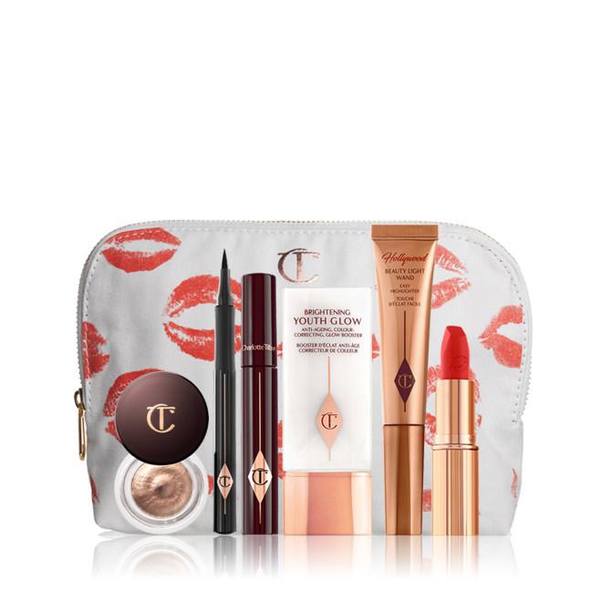 A light golden-coloured highlighter wand, mascara in dark crimson packaging, an open eyeliner pen, a primer in white and gold packaging, an open lipstick in a bright orange-red shade, and cream eyeshadow in an open glass pot. 