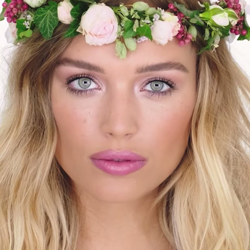 A fair-tone model with blue eyes wearing glowy, nude pink makeup with bright pink, matte lipstick along with a flower crown on her head. 
