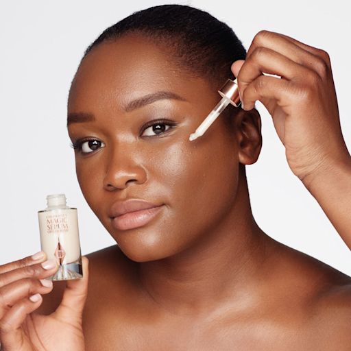 Magic Serum application on deep-tone model A deep-tone model with brown eyes wearing subtle eye makeup and applying a luminous face serum on her face with a gold-coloured dropper.