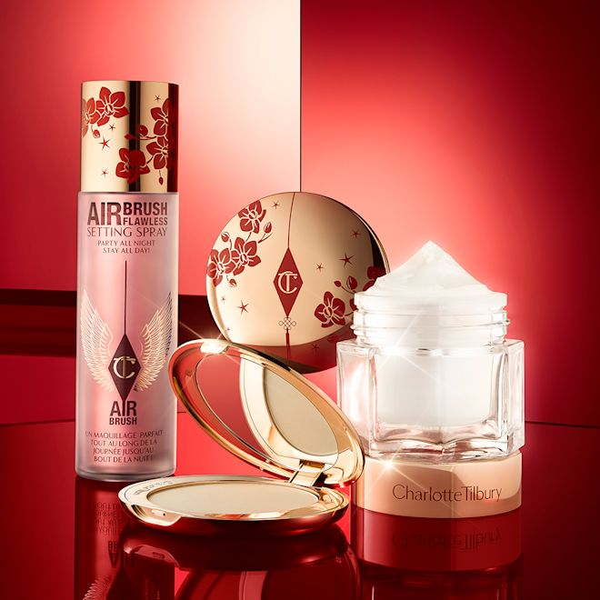 Lucky New Year Beauty Secrets displayed A matte burnt-orange red lipstick in gold-coloured packaging with cherry blossoms illustrated on the tube for the Lunar New Year, along with its lid next to it and an open, pressed powder compact for fair skin tones with a mirrored lid, in gold-coloured packaging with red-coloured cherry blossoms on the lid, setting spray with cherry blossoms on the lid, and a face cream in a glass jar with cherry blossoms on the lid.