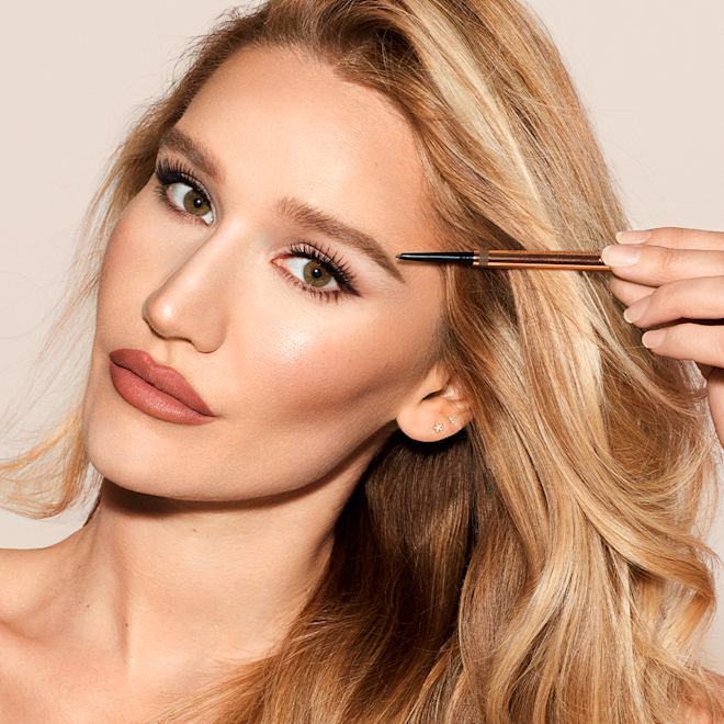 Sofia Tilbury Holding Brow Cheat Eyebrow Pencil Sofia Tilbury wearing matte, muted red lipstick with flawless and smooth bronzed makeup while filling in her eyebrows with an eyebrow pencil.