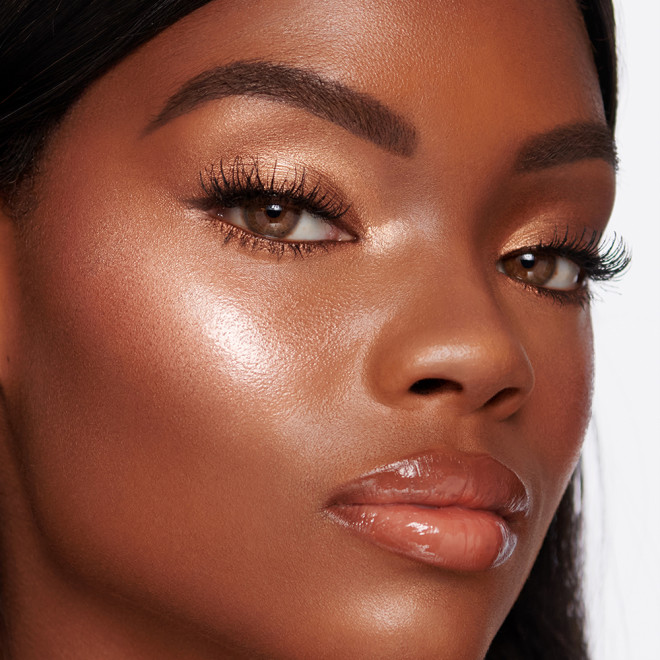 Deep-tone brunette model wearing shimmery bronze and gold eye makeup with glossy peach lipstick, and glowy medium-pink highlighter blush and candlelight-gold liquid highlighter. 