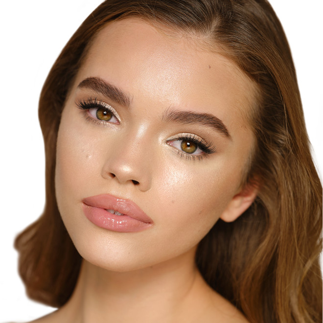 Medium-tone brunette model wearing glowy highlighter in a champagne-gold shade with nude pink lip lipstick and soft eye makeup.