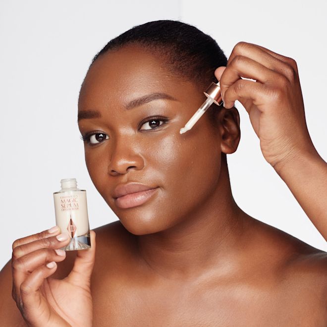 Magic Serum application on deep-tone model A deep-tone model with brown eyes wearing subtle eye makeup and applying a luminous face serum on her face with a gold-coloured dropper.