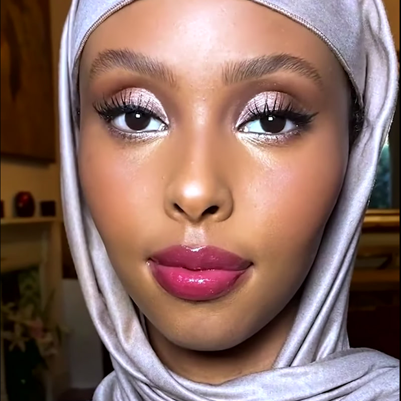 Deep-tone model with brown eyes and head-covering wearing shimmery lilac and smokey brown eye makeup with volumising mascara, black eyeliner, and bright magenta lipstick topped with lip gloss. 
