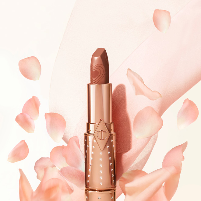 Banner with a lipstick with its lid removed in a nude peach shade in a gold-coloured tube with peach-coloured petals falling on it.