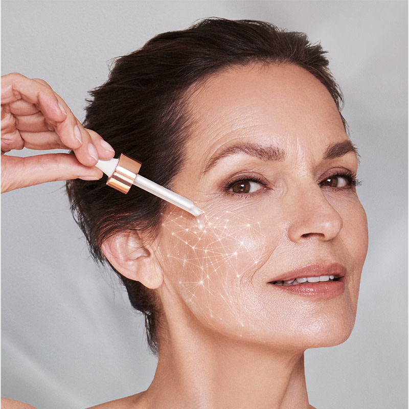 A light-tone model with mature skin applying a luminous, ivory-coloured face serum with a dropper. 