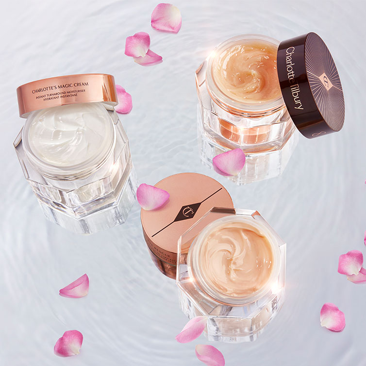 Three open, glass jars with pearly-white face cream, peach-coloured, thick night cream, and champagne-coloured eye cream with their lids next to them, and all three placed on crystal-clear water with rose petals surrounding them.