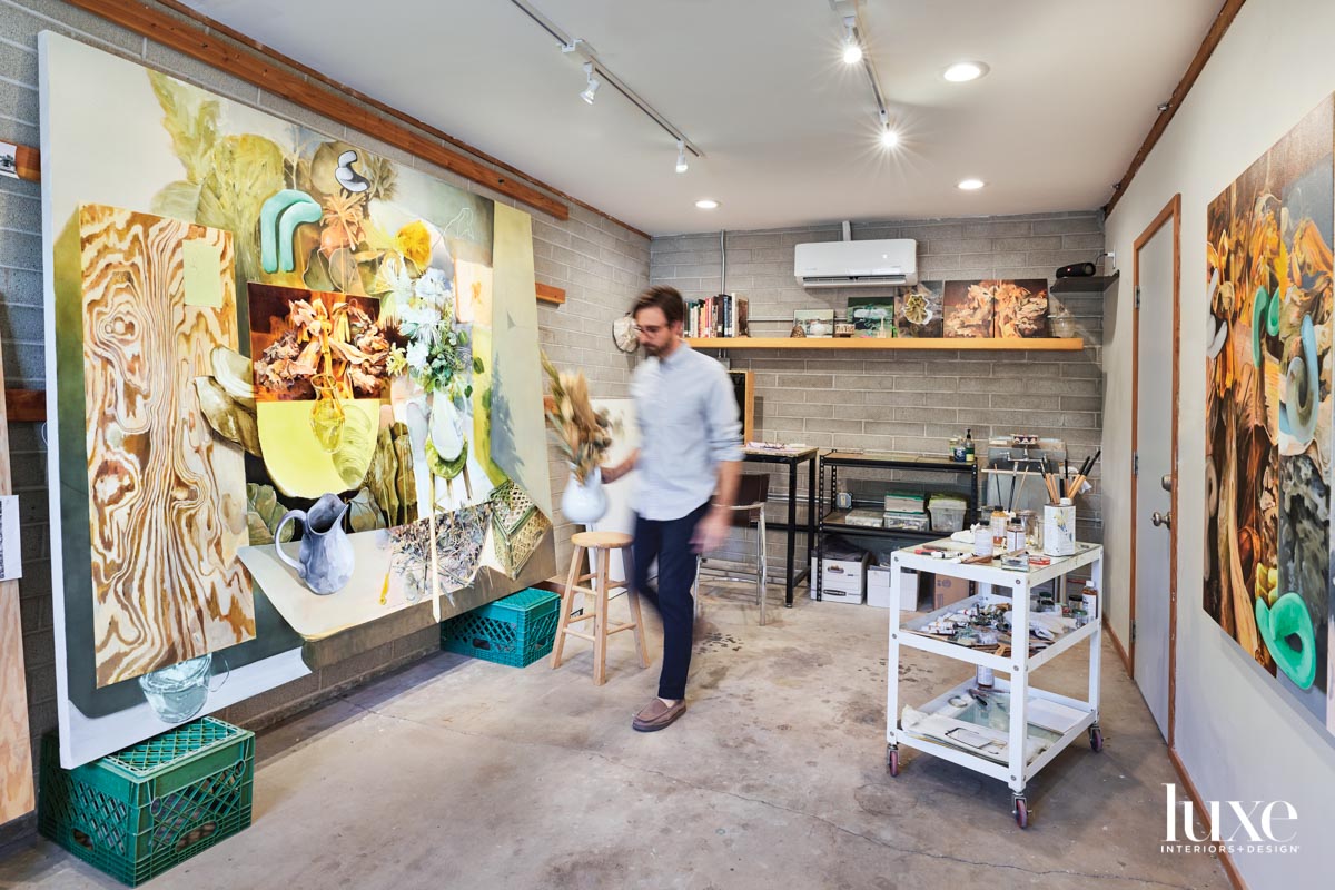 A man walking through a studio holding a vase with flowers. Next to him is a large collaged painting.