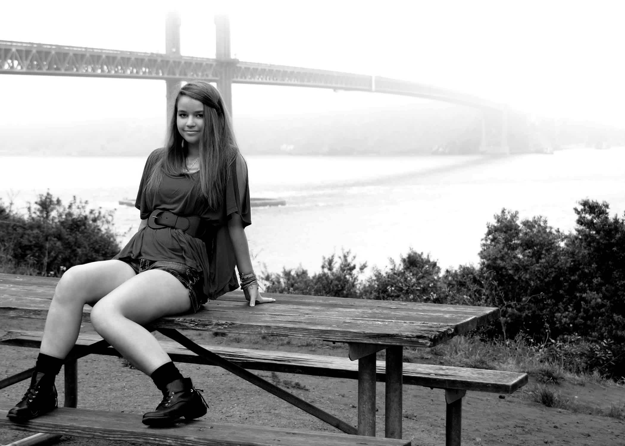Girl sitting on a picnic table in front of a bridge.
