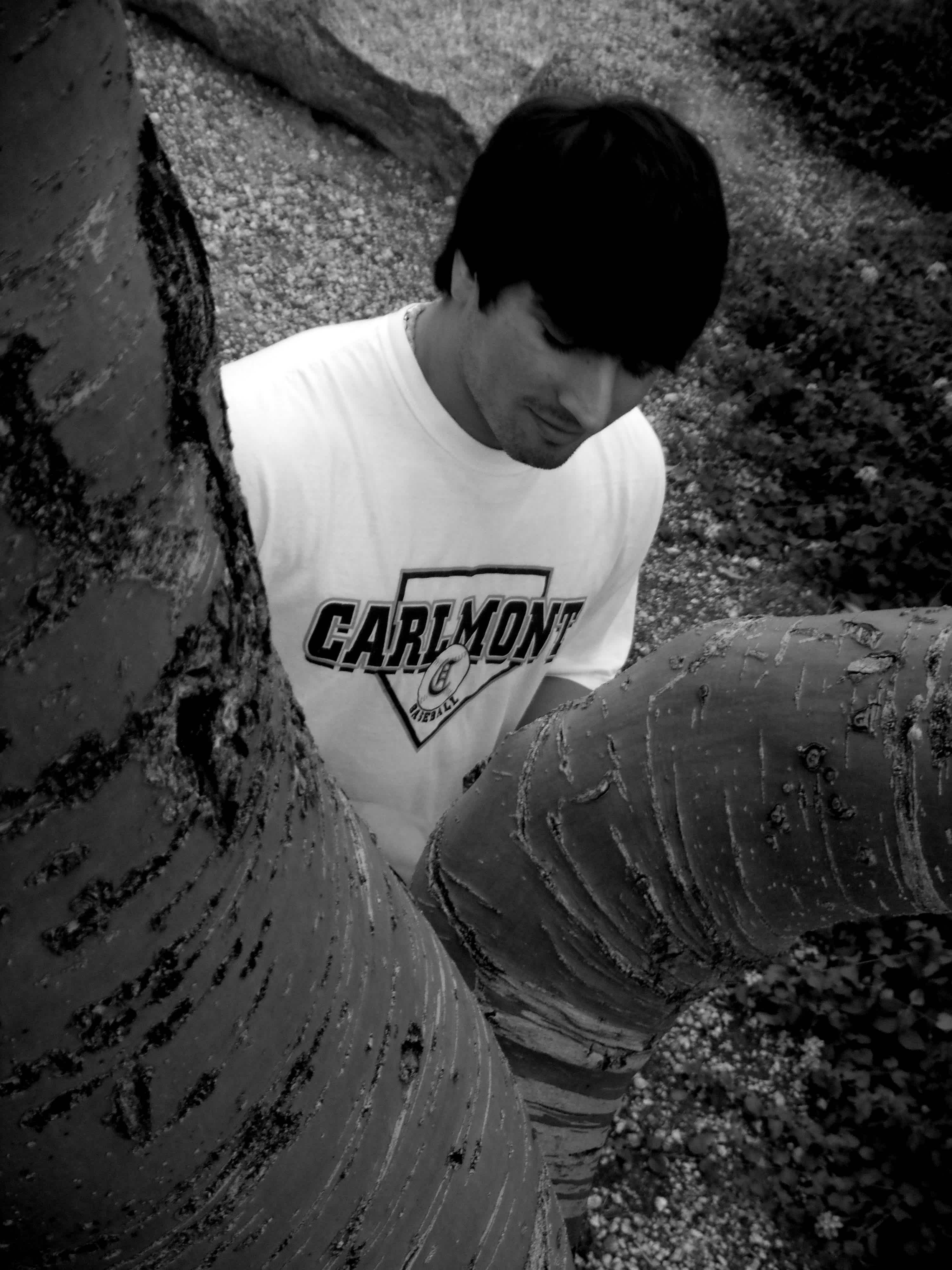 Black and white boy in Carlmont t-shirt standing behind the trunk of a tree. 