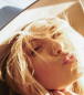 Close up image of a woman's face with sun shining on it. She is wearing a hat, has light blond hair, and is making a "kissing" face.