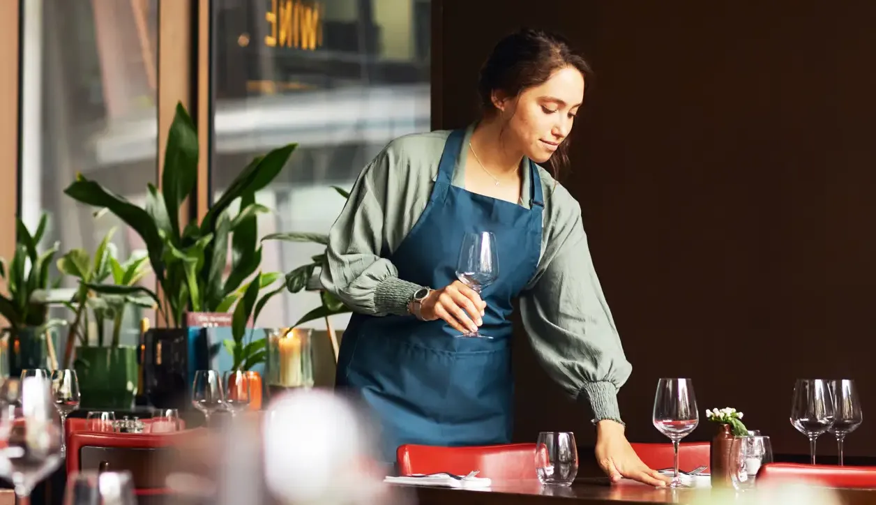 Woman in a blue apron holding an empty wine glass in her hand. She's got the other hand on a table in a bar which has more empty glasses on it.