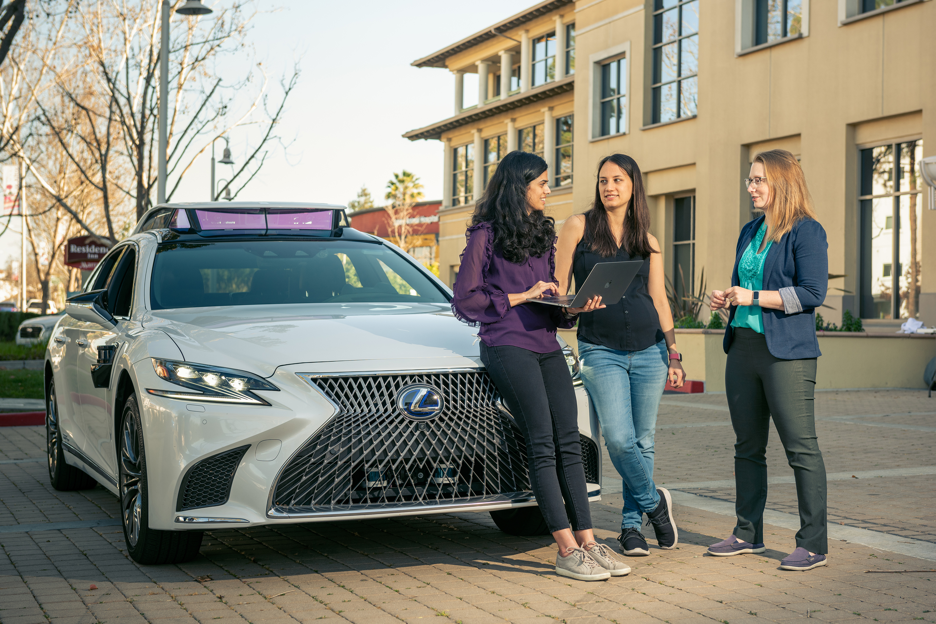 Three women standing next to a Lexus. One of them is holding a laptop.