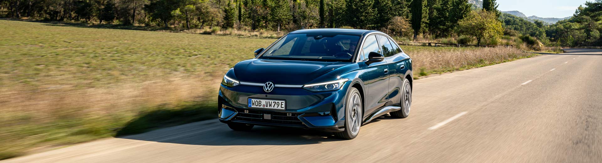En-mörkblå-Volkswagen-ID.7-Tourer-elkombi-kör-på-en-asfalterad-väg-genom-ett-landskap-med-fält-och-träd