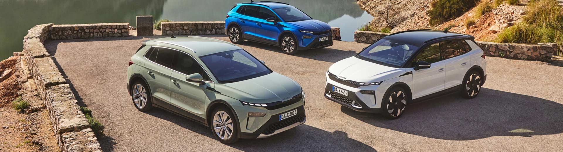 Tre-stycken-Skoda-Elroq-elbilar-i-färgerna-mintgrön,-vit-och-blå-uppställda-på-en-utsiktsplats-vid-en-sjö-och-bergigt-landskap
