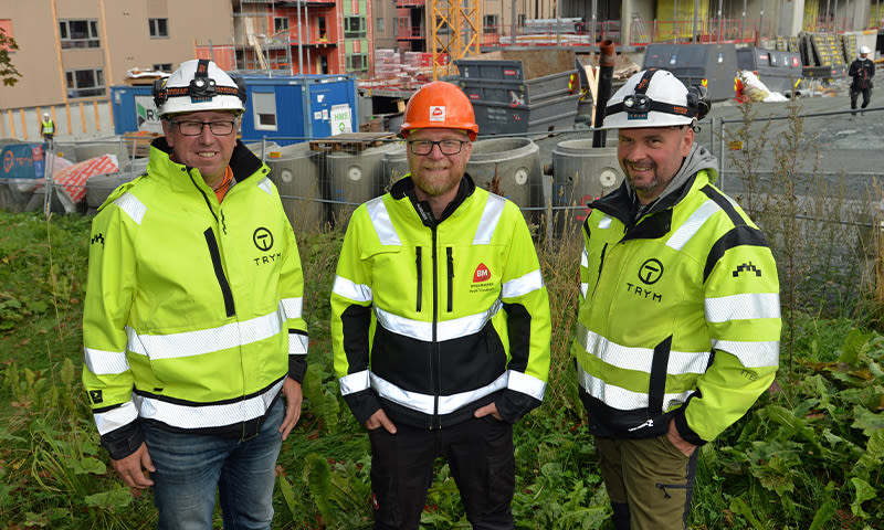 Tre byggearbeidere i neongule refleksjakker og hjelmer foran byggeplass med stillas og containere