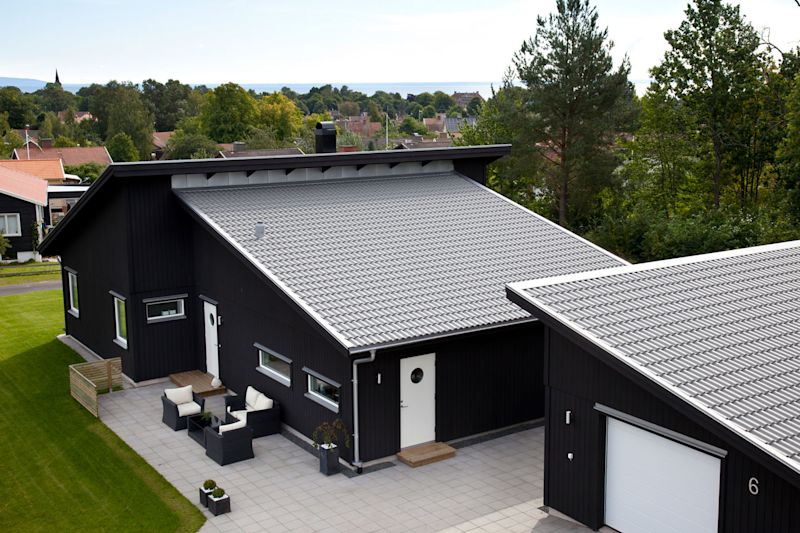 Moderne svart hus med skrå grått tak, hvit dør, uteplass med sittegruppe og grønn plen