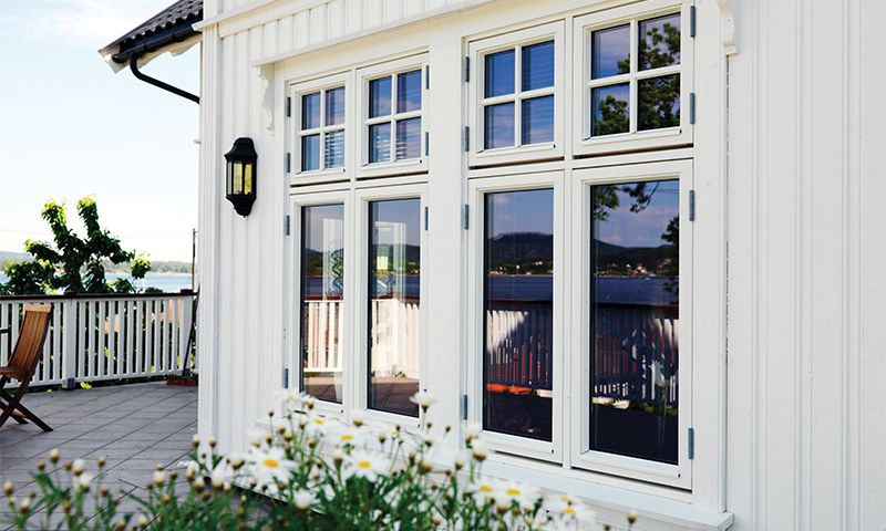 Hvitt trehus med store vinduer, blomstrende prestekrager og terrasse mot blå innsjø.