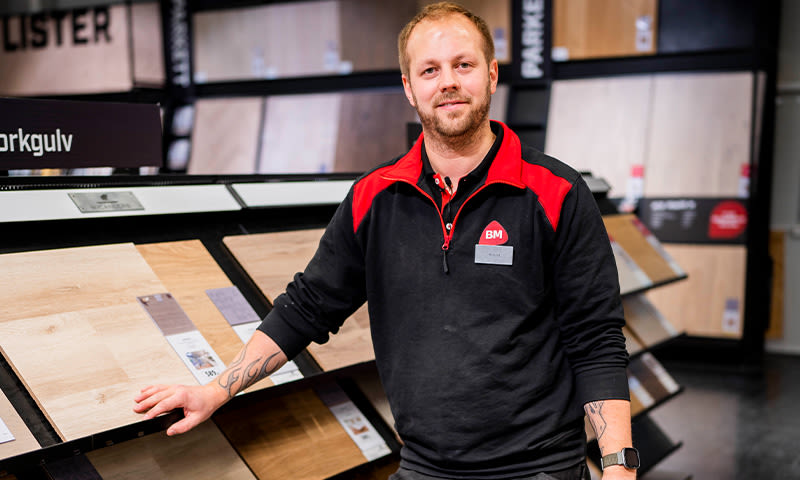 Ansatt i rød-svart BM-uniform ved utstilling av lys og mørk parkettgulv i butikk.