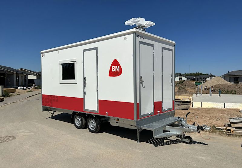 Hvit og rød to-akslet byggebrakke med BM-logo, parkert på byggeplass under klar blå himmel.