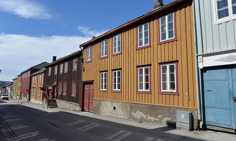 Rad av gule, brune, røde og lyseblå trehus langs stille gate under blå himmel.