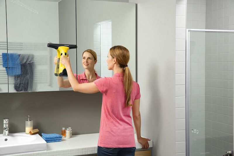 Person i rosa t-skjorte pusser et speil med gul vindusrengjører på hvitt baderom med blå håndklær.