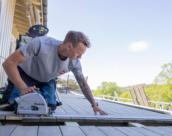 Håndverker i grå t-skjorte sager lyst terrassebord med hvit Makita-sag under blå himmel.