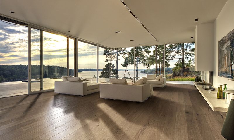 Moderne stue med beige sofaer, mørkt tregulv og store vinduer mot innsjø og skog