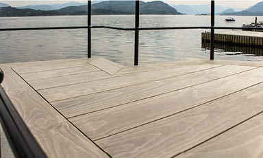 Lysebrun trebrygge ved fjord med svart rekkverk, båt og fjell i bakgrunnen.