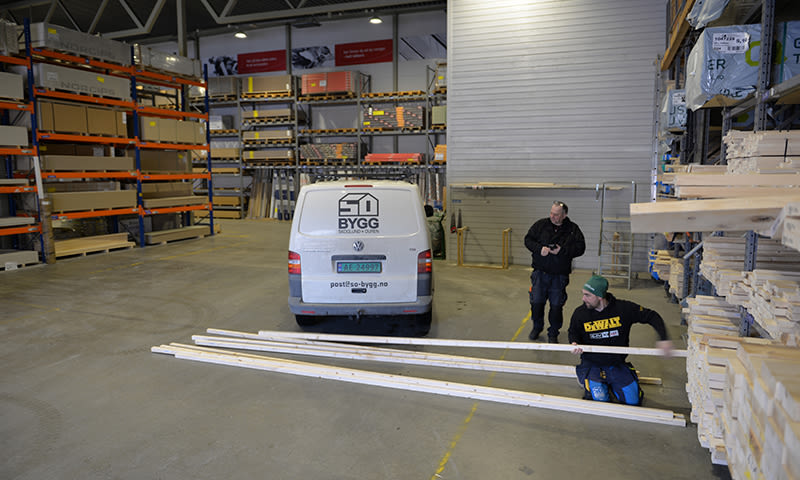Lyst byggevarelager med hvit BYGG-varebil, to håndverkere måler trelekter ved stabler.