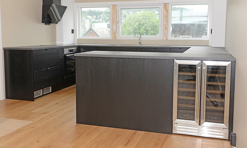 Mørktgrått L-formet kjøkken med sort benkeplate, innebygd vinkjøler og vindu mot grønn utsikt.