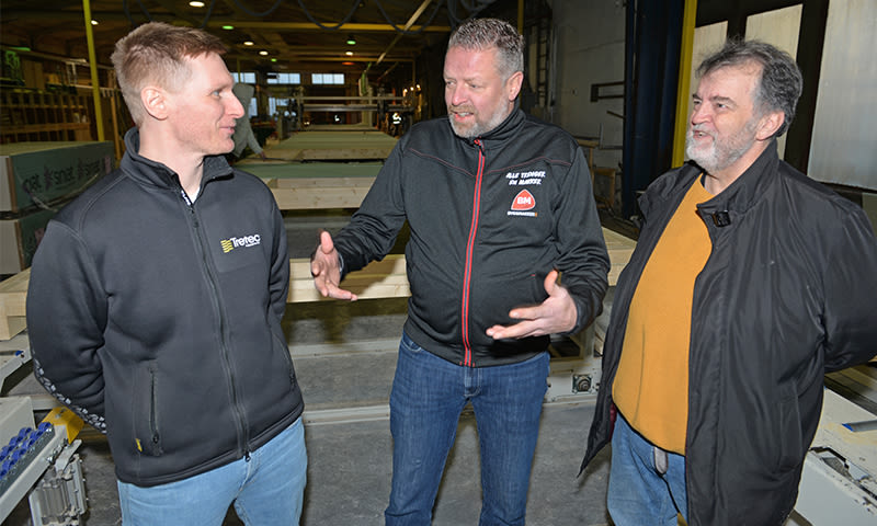 Tre menn i verkstedmiljø ved transportbånd med treplater, iført sorte jakker og jeans.