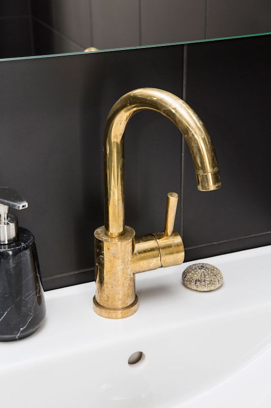 Gyllen tappekran på hvit vask ved svart flisvegg, marmor såpedispenser og sjøpiggskall.